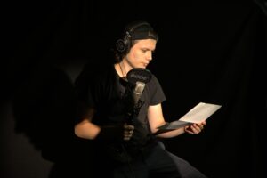 Cette image montre une personne en train d'enregistrer un audio ou une voix off dans un studio sombre. La personne porte un casque audio et est assise devant un microphone de marque RØDE. Elle tient une feuille de papier, probablement un script, qu'elle lit tout en enregistrant. L'éclairage est concentré principalement sur son visage et ses mains, créant un effet de clair-obscur qui met en avant l'ambiance de travail en studio. La personne est habillée en noir, ce qui se fond dans le fond sombre, renforçant l'atmosphère focalisée et professionnelle de la scène.