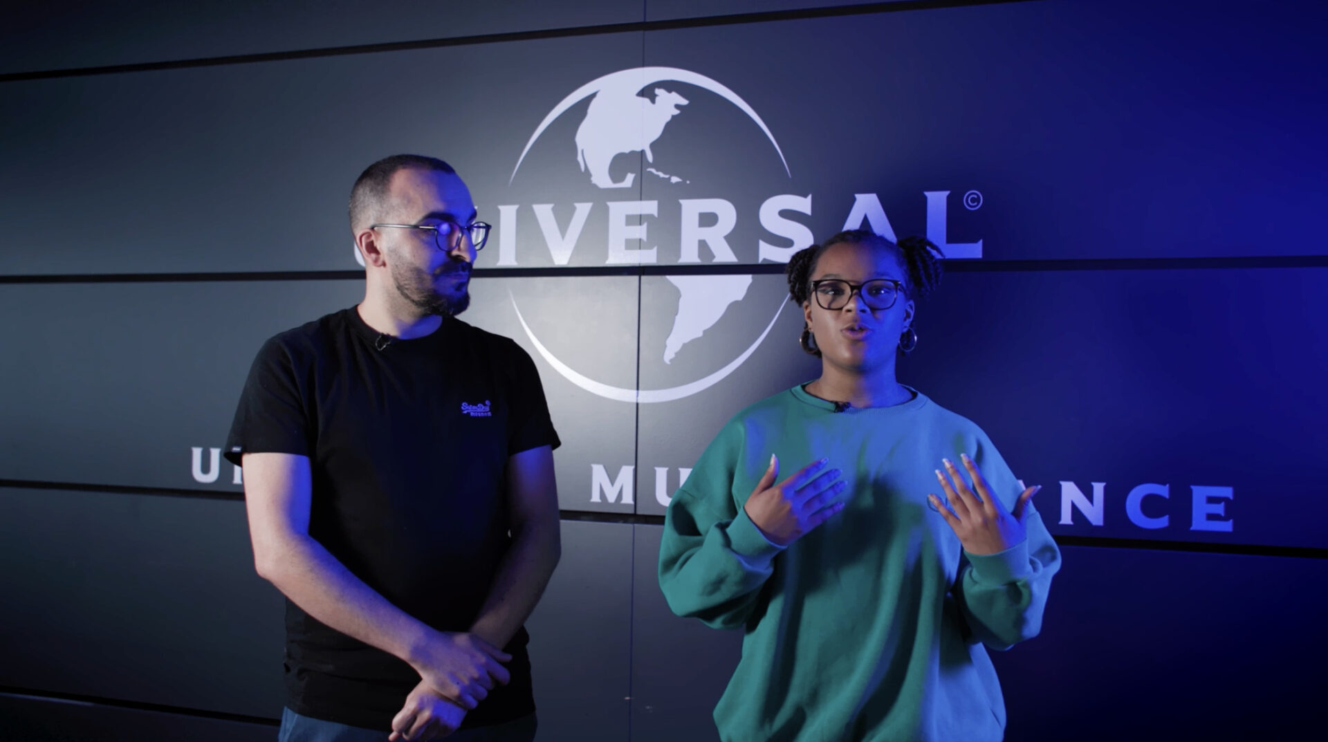 Cette image montre deux personnes debout devant un logo Universal Music, qui est visible en arrière-plan. La scène semble se dérouler dans un cadre professionnel, probablement au sein d'un bureau ou d'un studio appartenant à Universal Music. Les deux personnes portent des lunettes et sont éclairées par un éclairage bleu. La personne à gauche est habillée en noir, tandis que celle à droite porte un pull vert. Ils semblent être en pleine discussion ou présentation, la personne à droite gesticulant pour accompagner son discours.