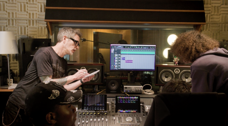 Un groupe de personnes collabore dans un studio d'enregistrement, entouré d'équipements professionnels tels qu'une console de mixage, des écrans et des enceintes. Un homme aux cheveux gris prend des notes, tandis que l'écran principal affiche un logiciel de production audio avec des pistes en cours d'édition.