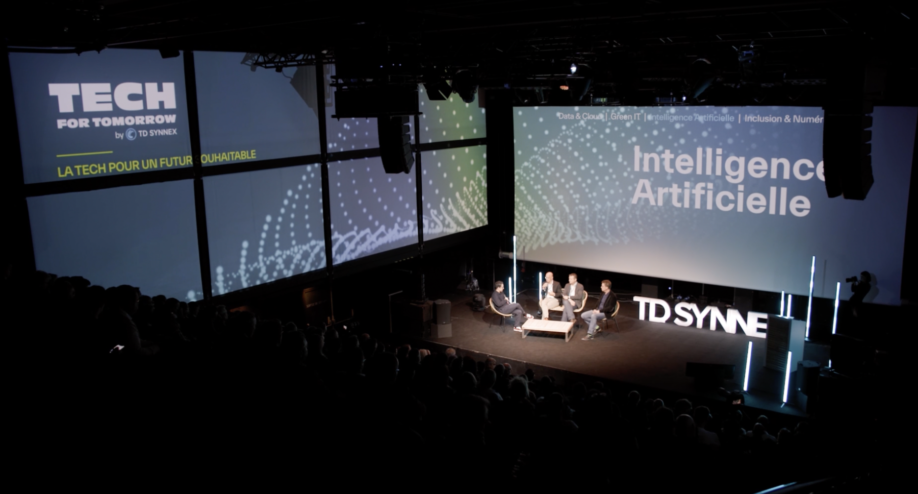 Une conférence sur l'intelligence artificielle organisée par TD SYNNEX, avec quatre intervenants discutant sur scène dans une salle sombre. Les écrans géants affichent les mots 'Tech for Tomorrow' et 'Intelligence Artificielle' en arrière-plan, sous le slogan 'La tech pour un futur souhaitable'.