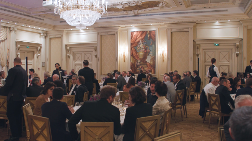 Un dîner formel dans une salle de réception élégante avec des invités assis à des tables rondes. La pièce est ornée de lustres, de moulures et de tableaux, avec des serveurs en uniforme circulant parmi les convives.