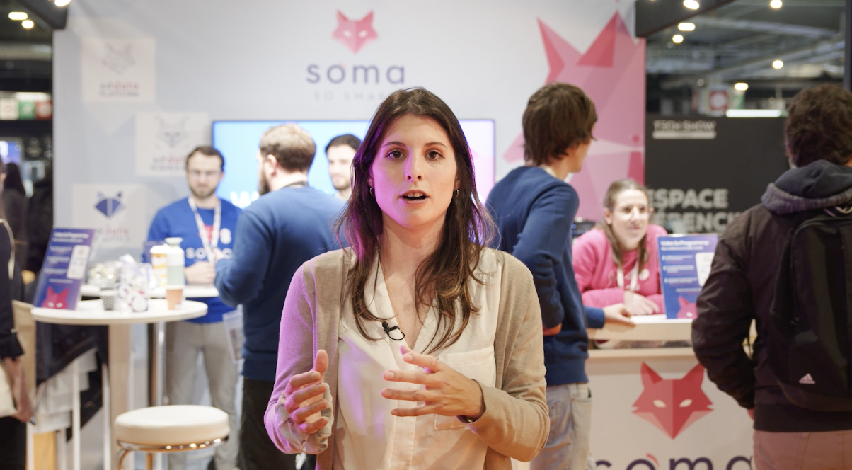 Une femme est en train de parler face caméra lors d'un événement professionnel. En arrière-plan, plusieurs personnes discutent devant un stand de l'entreprise "Soma".