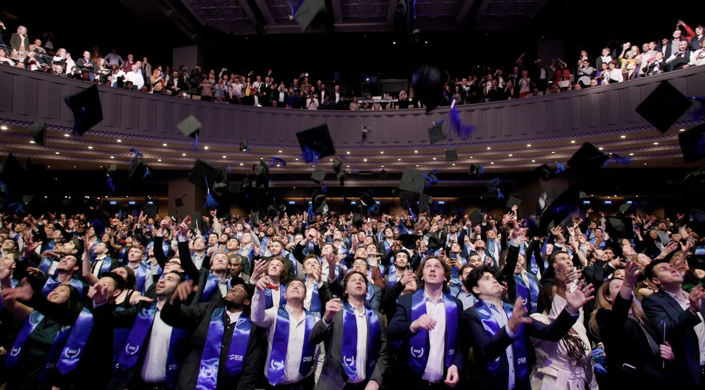 Des étudiants en tenue de graduation célèbrent en lançant leurs toques en l'air dans un auditorium bondé. La foule est joyeuse et des spectateurs les applaudissent depuis les balcons.