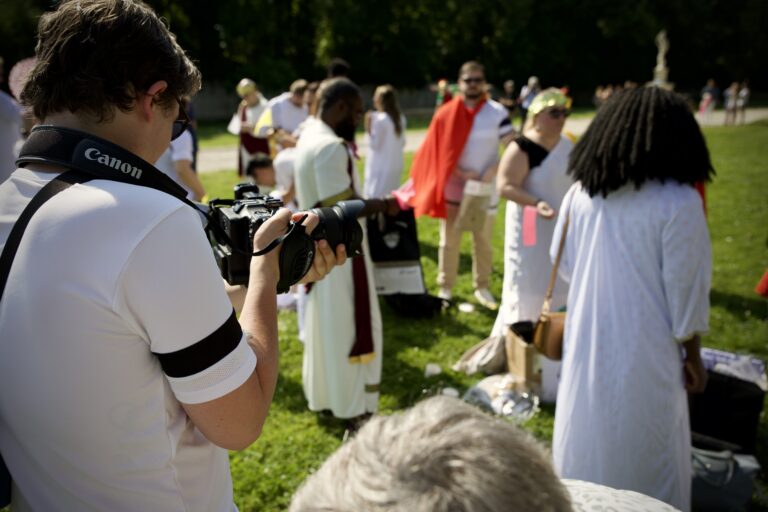 Cameraman de Graine de Production capturant les moments du teambuilding romain de CPMS, avec des participants en tenues romaines en arrière-plan.