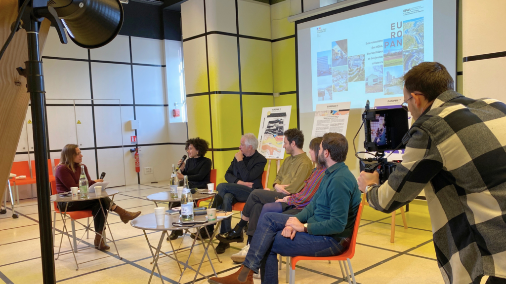 Un caméraman de Graine de Production filme une table ronde lors d'une conférence Europan 17. Les intervenants, assis devant un écran de présentation, discutent dans une salle aux murs jaunes et noirs.