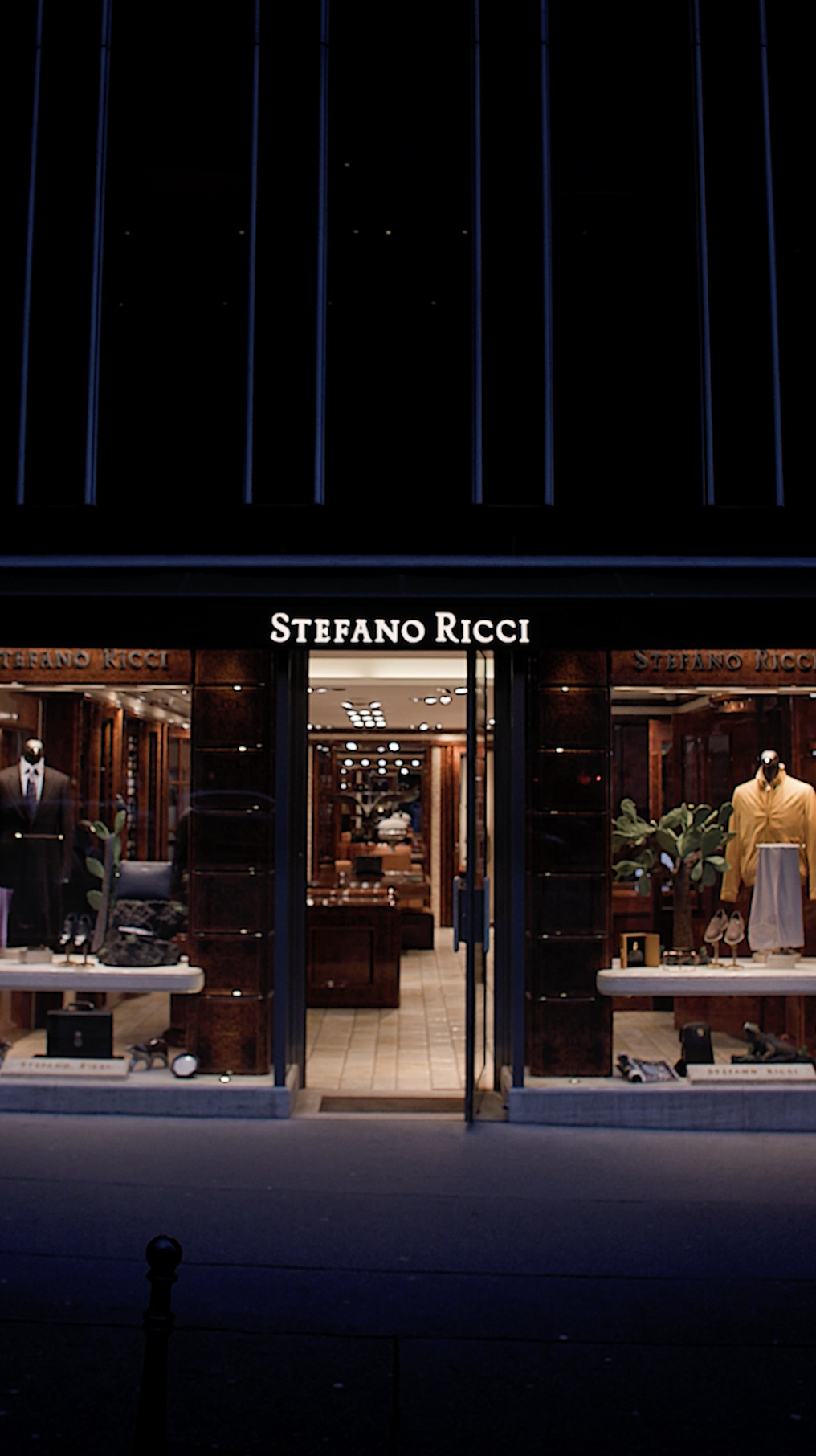 L'image montre la devanture d'une boutique de luxe "Stefano Ricci". La façade est élégante avec une grande porte d'entrée vitrée, encadrée par des vitrines bien éclairées. À l'intérieur, on peut voir des vêtements de haute qualité, probablement des costumes, et divers accessoires soigneusement présentés. La boutique donne une impression de raffinement et de sophistication, caractéristique des marques de luxe italiennes. L'éclairage doux et la disposition des articles créent une atmosphère accueillante et exclusive.