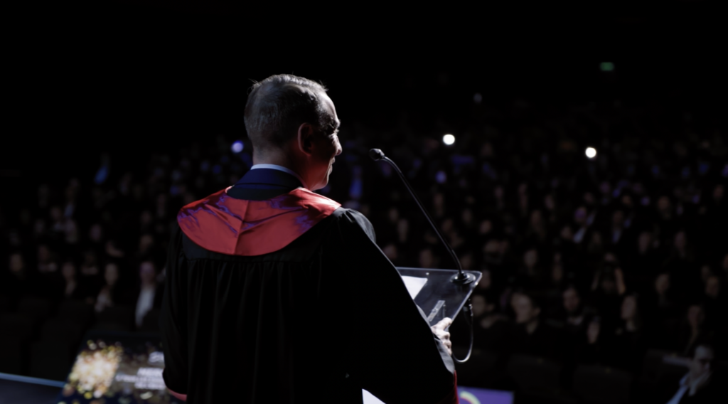 Un orateur en toge académique, vu de dos, s'adresse à un public nombreux lors d'une cérémonie de remise de diplômes. Le micro et la lumière centrée sur lui contrastent avec la salle sombre remplie d'invités lors de la remise des diplômes de l'ESCE.