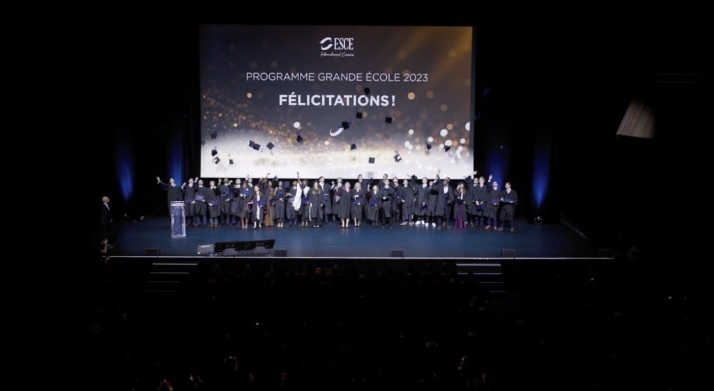 Des diplômés en toges et chapeaux de graduation célèbrent sur scène, levant leurs mains et diplômes vers le ciel face à une audience enthousiaste. Un conférencier en robe rouge se tient au centre, levant les bras vers le public sous un projecteur lumineux à l'occasion de la remise des diplômes ESCE.