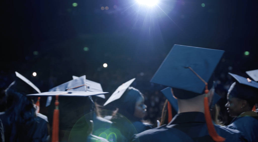 Des diplômés en toges et coiffés de toques se tiennent sur scène, éclairés par un projecteur brillant dans une salle sombre remplie de spectateurs. L’ambiance met en valeur la solennité et l’émotion de la cérémonie de remise des diplômes.